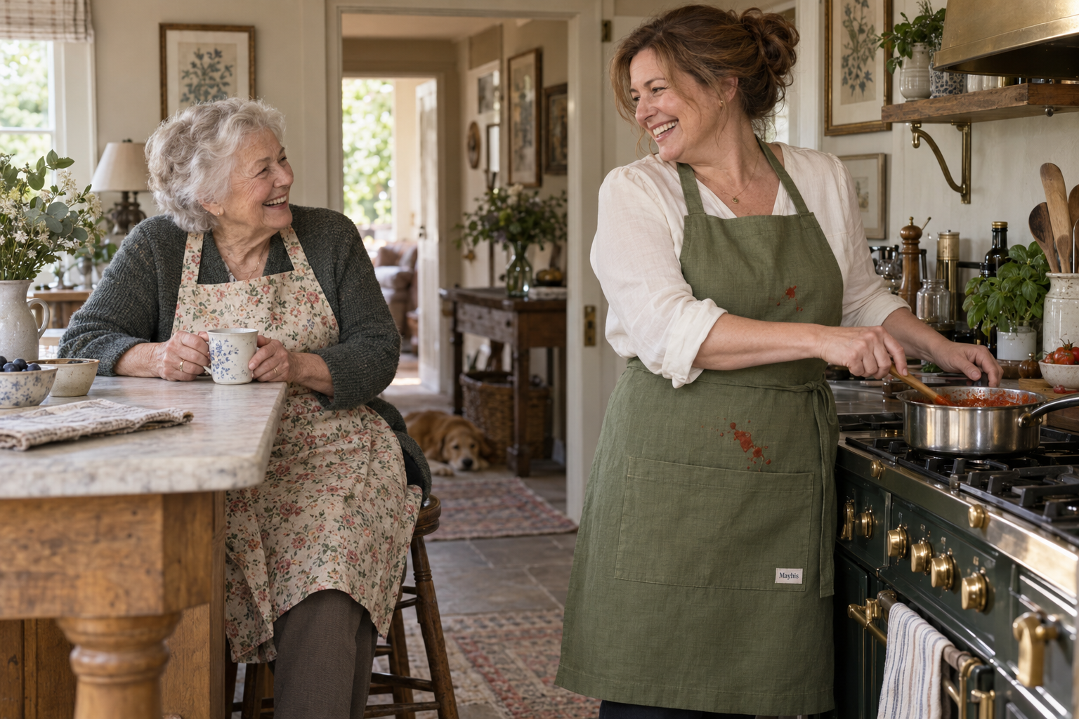A mother in a floral apron shares a laugh over coffee with her daughter, whose olive Maybis apron has caught a splash of tomato sauce while cooking. A dog rests in the background.